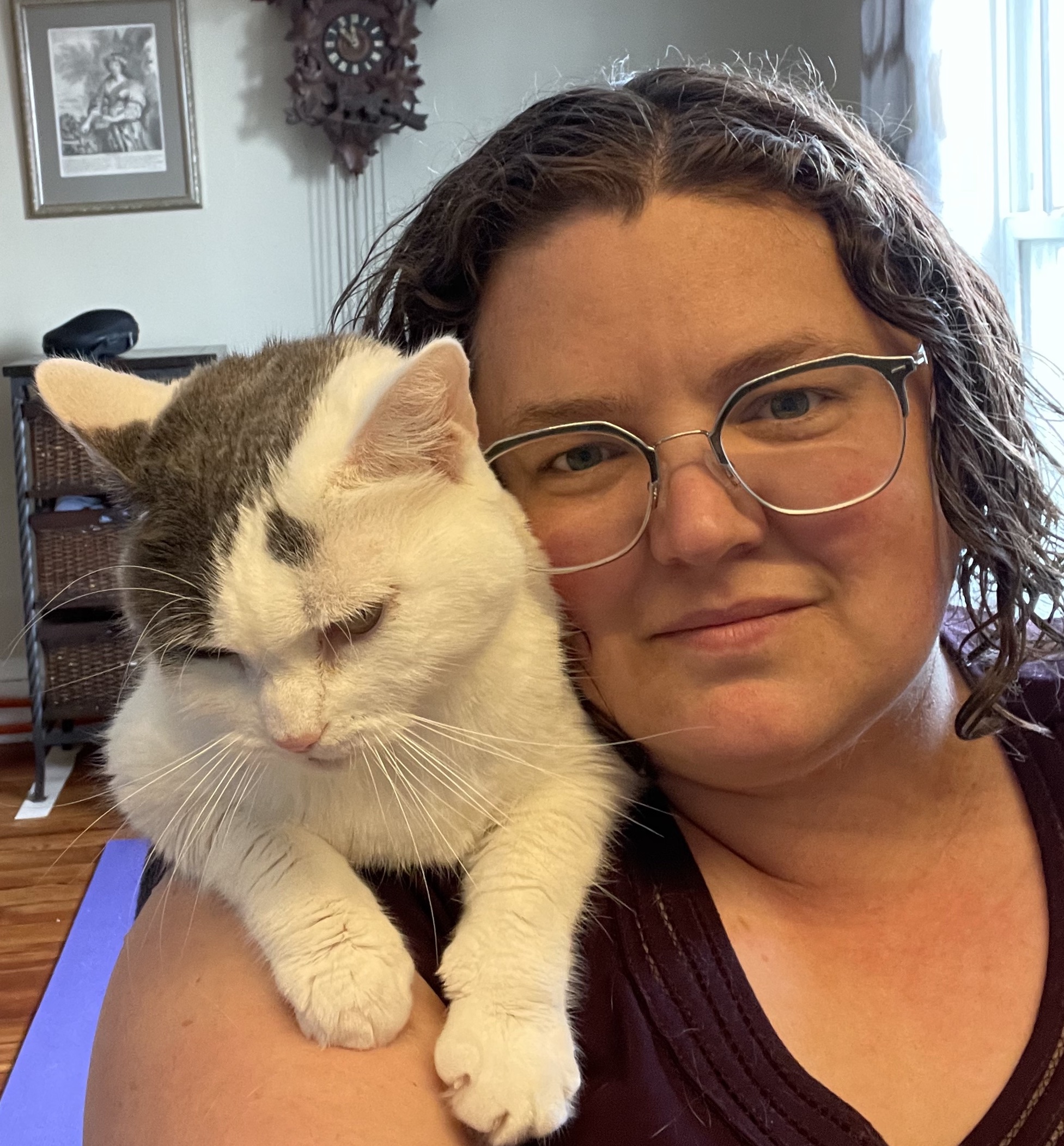A white woman looks straight at the camera with a half-smile. She has curly brown hair and glasses. There's a soft white-and-grey cat draped over her shoulder.