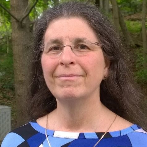 A white woman looks straight at the camera with a half-smile. She has curly brown hair and glasses. There's a soft white-and-grey cat draped over her shoulder.