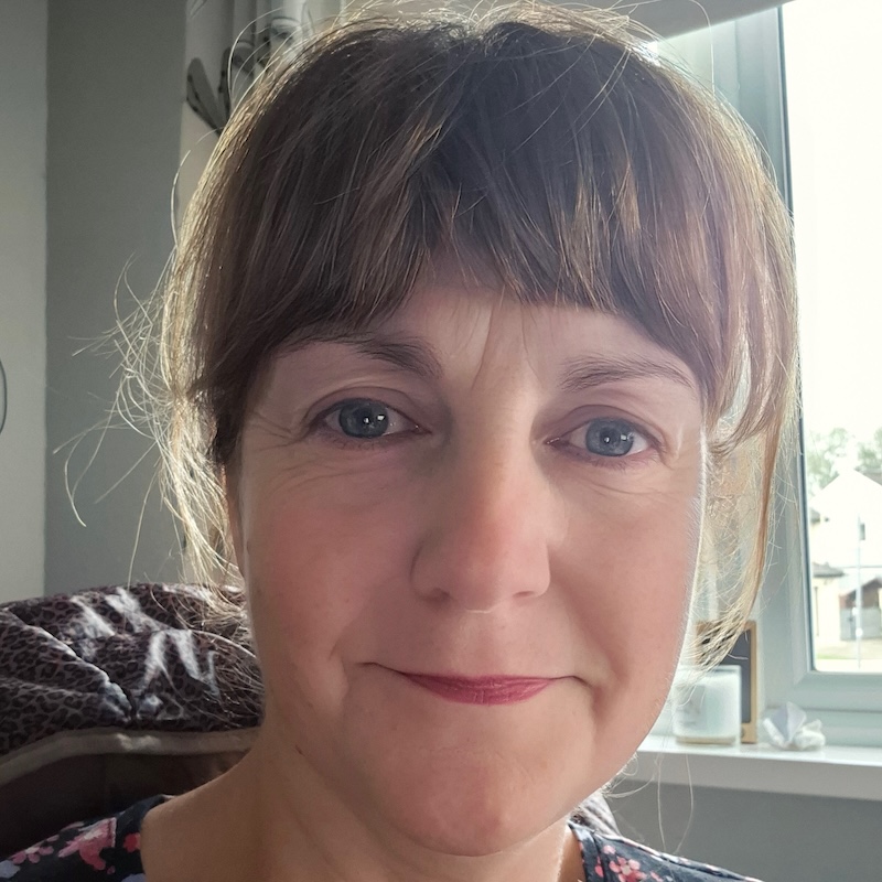 A white female with brunette hair tied back and a fringe needing a brush, wearing a floral top by a window, smiling while working from home as she enjoys the thought of the weekend ahead and the joy of no long commute.