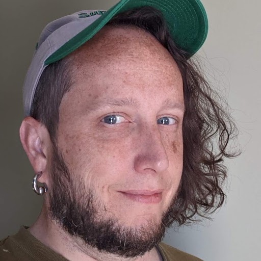 A white, freckle-faced male with blue eyes, chin-length curly dark brown hair, and a beard is smiling in front of a light green wall. He is wearing a green t-shirt and a grey & green Seattle Supersonics baseball cap.
