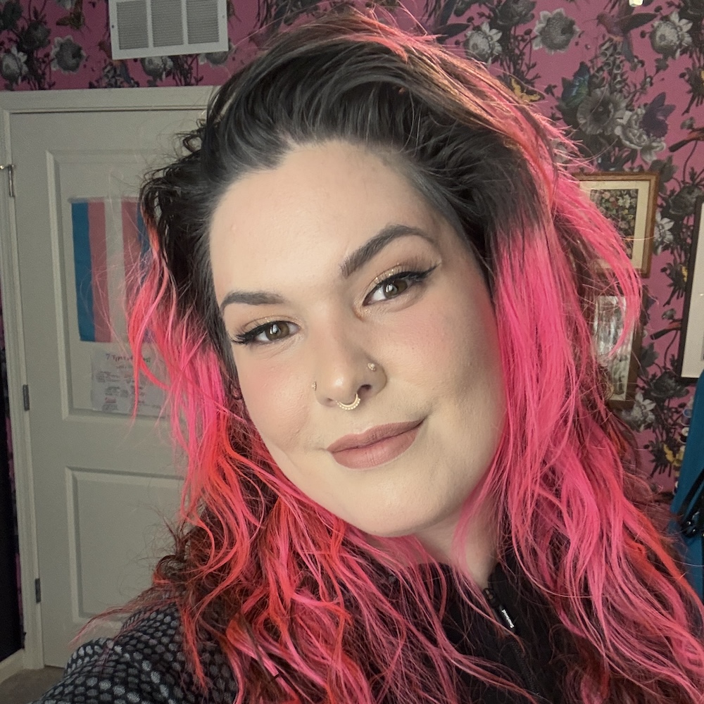 A woman with a friendly smile. She has light skin, hazel eyes, and pink highlights in her dark hair. She is wearing a dark colored outfit and has facial piercings on her nose.