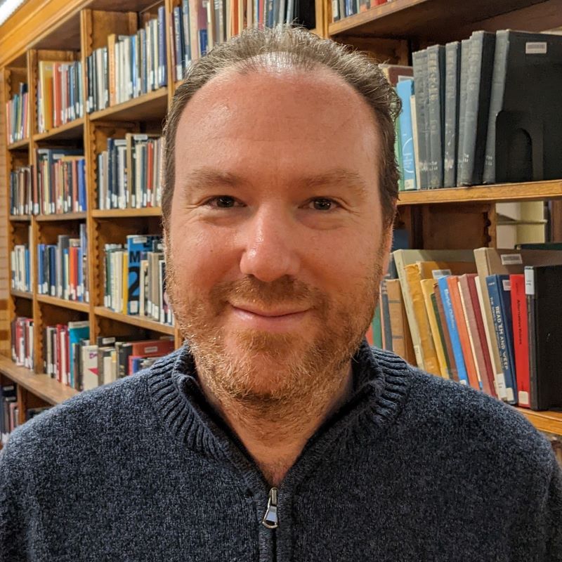 Headshot of a white man standing in front of library stacks. He has short curly hair and a short beard, both of which still probably need a trim.