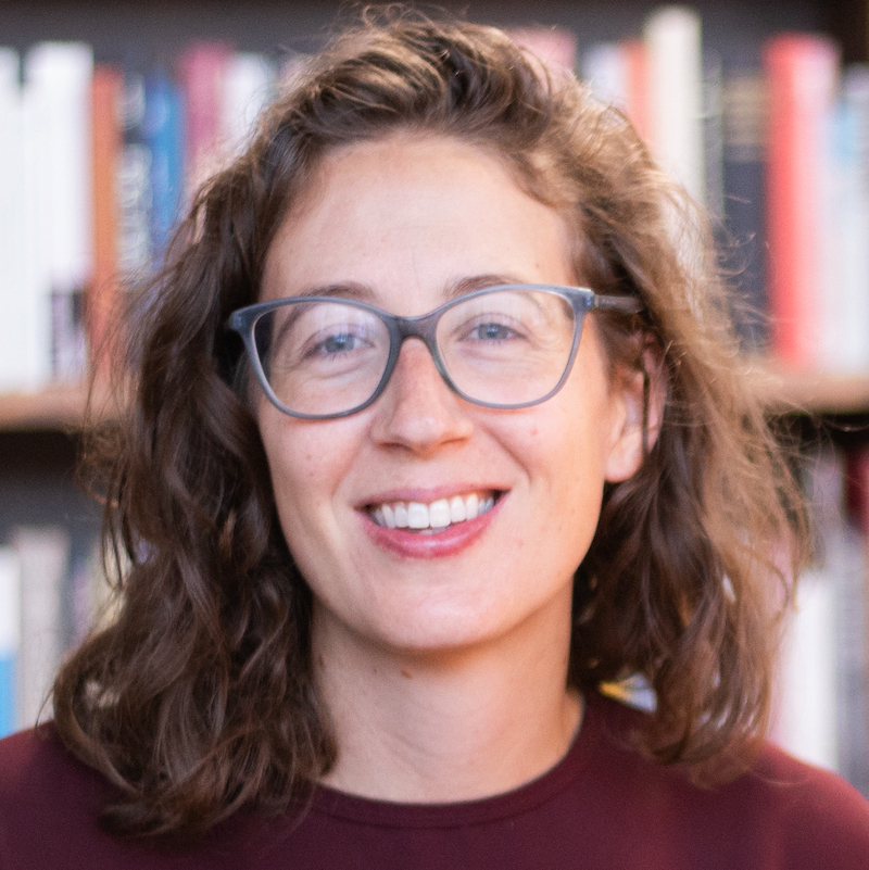 Close up white woman with brown hair and light blue glasses, with library books in background.