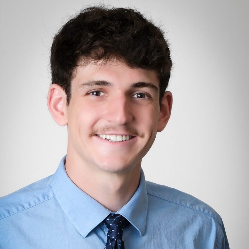  A young man with dark, wavy hair and a mustache smiling in a professional headshot. He is wearing a light blue button-down shirt and a dark navy blue patterned tie against a neutral, light gray background.