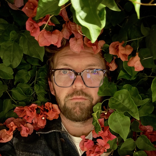 Headshot of middle aged white man with a beard and glasses being gently engulfed by flowers and bushes.