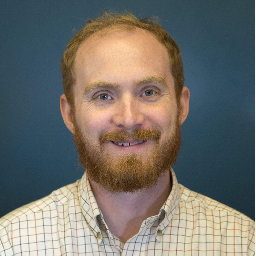 A white man with a short reddish beard and scant tuft of hair stares at the camera wearing a white collared shirt against a dark blue background.