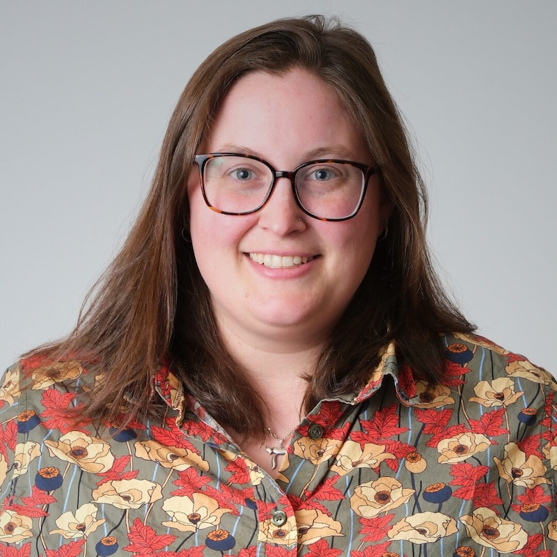 A white woman with shoulder-length brown hair and glasses faces the camera in a professional headshot. She wears a floral button-up shirt against a soft light gray studio background.