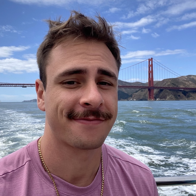 A close-up selfie of Kyle Reiley outdoors on a boat, with the Golden Gate Bridge visible behind him. He has short brown hair that is slightly wind-tousled, thick dark eyebrows, and a prominent brown mustache. His expression is relaxed and faintly smiling, with a calm, content look. He is wearing a dusty pink short-sleeve T-shirt and a gold chain necklace that rests against his collarbone. Behind him, the water is choppy and deep blue-green, with white foam trailing behind the moving boat. In the distance, the red-orange towers and suspension cables of the Golden Gate Bridge stretch across the frame, partially framed by rolling brown and green hills. The sky is bright blue with scattered wispy white clouds, suggesting a clear, breezy day. The overall scene conveys motion from the boat, cool coastal air, and a scenic view of San Francisco Bay.