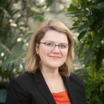 Maureen Cresci Callahan, a white woman with blond hair and glasses wearing an orange dress and black blazer. Greenery is in the background. She has a calm expression.