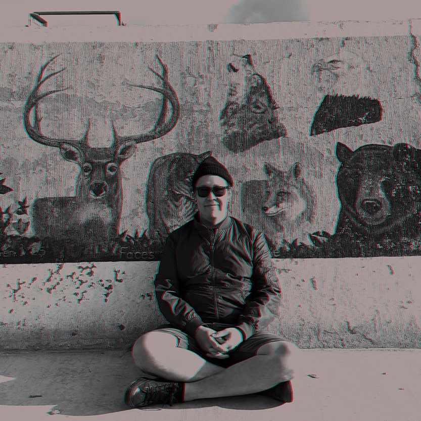 A person sits comfortably cross-legged before a mural of animals, bundled in a jacket and beanie, projecting a reflective mood with a hint of quiet humor.