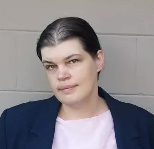 Close up of a white person in a suit coat against a cinderblock wall.